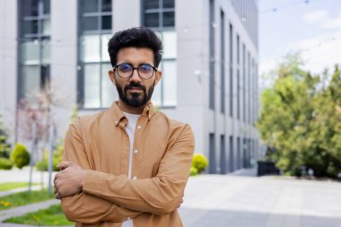 Ciddi düşünen iş adamı kameraya çapraz kollu bakıyor, Hintli adam ofis binasının dışında gömlekli..