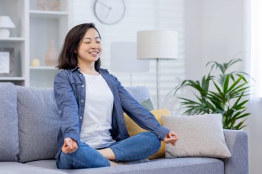 Genç gülümseyen Asyalı kadın, oturma odasında kanepede meditasyon yapıyor, gözleri kapalı sevinçle gülümseyen kadın, gelecekteki planları düşünüyor ve hayal ediyor, başarının hayalini kuruyor..