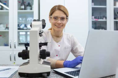 Laboratuvarda çalışan genç bir kadın doktorun portresi, elinde mikroskopla bir masada oturuyor ve bir not defterine yazıyor, gülümsüyor ve kameraya bakıyor..