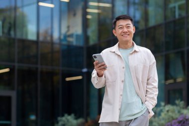 Genç Asyalı iş adamı, serbest çalışan, tasarımcı ofis binalarının önünde cep telefonu kullanıyor ve kameraya gülümsüyor..
