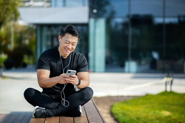 Asian young male sportsman sitting on bench in lotus position near lake in park wearing headphones and listening to music, podcast, talking on phone. Resting.