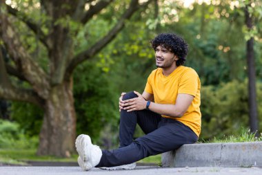 Genç Hintli bir adam parkta spor yapıyor, sabah yürüyüşüne çıkıyor. Bacağından yaralanmış, dizini tutuyor, ağrısı var..