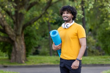 Parkta parka oturmuş elinde spor minderiyle bekleyen Hintli genç bir adamın portresi. Spor, spor, yoga..
