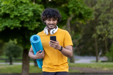 Genç aktif Hintli adam parkta duruyor kulaklık takıyor, spor minderi tutuyor ve telefon kullanıyor..