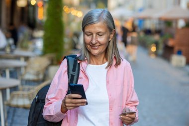 Akşam vakti sırt çantası ve telefonla gezen yaşlı, olgun bir bayan turist, gri saçlı, emekli bir kadın elinde banka kartı ile mutlu bir şekilde gülümsüyor ve online alışveriş için akıllı telefon uygulaması kullanıyor..