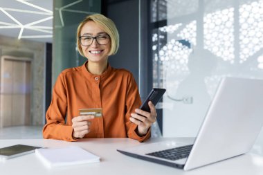 İş yerindeki genç ve güzel bir kadının portresi, elinde telefon ve banka kartı olan bir iş kadını gülümseyen ve kameraya bakan bir kadın, online hizmetler ayarlayan ve malları satın alan bir kadın.