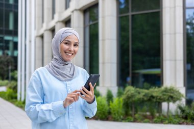 Başörtüsü içinde genç bir iş kadını ofis binasının dışından şehre giriyor. Kadın telefonda başvuru yapıyor, gülümsüyor ve kameraya bakıyor. Çevrimiçi arama için akıllı telefonu tutuyor..