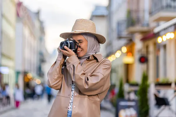 ヒジャーブの夜の街を歩く若い美しい女性 カメラと帽子をかぶる観光客は 旅行中のイスラム教徒の女性が満足して笑顔で歴史的な街を検査します — ストック写真