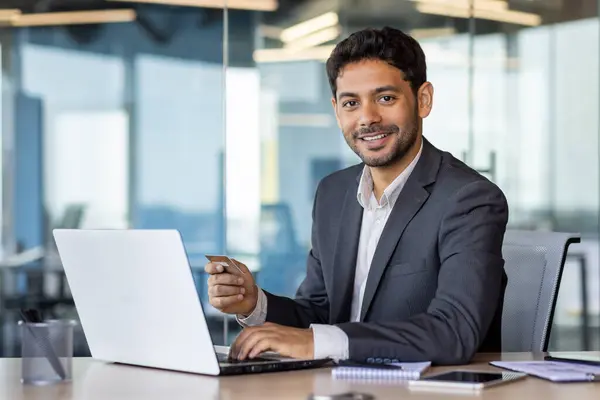 Portrait of young hispanic businessman inside office at workplace, man holding bank credit card in hands, using laptop for online shopping and money transfer, employee smiling and looking at camera.
