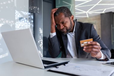 İş yerindeki üzgün ve endişeli işadamı, aldatan adam online para transferi hatası aldı, Afrikalı Amerikalı adam ofis içinde dizüstü bilgisayarında banka kartı tutmaktan memnun değil.