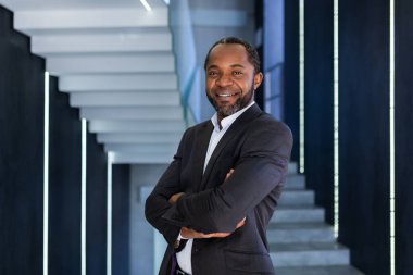 Ofis içinde olgun ve başarılı bir iş adamının portresi, Patron kollarını kavuşturup kameraya bakıyor, Afro-Amerikan takım elbiseli adam başarılı bir iş başarısı elde etmekten mutlu..