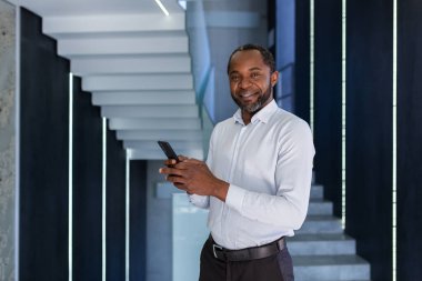 Olgun deneyimli Afro-Amerikan bir adam ofisin içinde elinde telefonla dolaşıyor gömlekli bir işadamı portresi, gülümsüyor ve akıllı telefondaki uygulamayı kullanarak kameraya bakıyor..