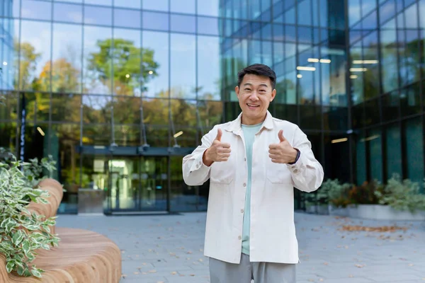 Asian businessman, founder of a firm and company standing in front of an office center, smiling at the camera and showing super success signs with his fingers.