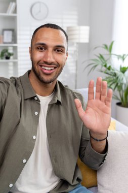 Dikey fotoğraf. Genç, gülümseyen Müslüman bir adam evinden online bir video yayını yapıyor. Kanepede oturup kameraya el sallamak..
