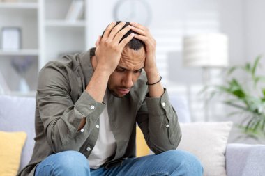Üzgün ve yorgun genç Afrikalı Amerikalı adam evdeki kanepede oturuyor, eğiliyor ve el ele tutuşuyor. Depresyon geçiriyor, panik atak geçiriyor, sorunları düşünüyor..