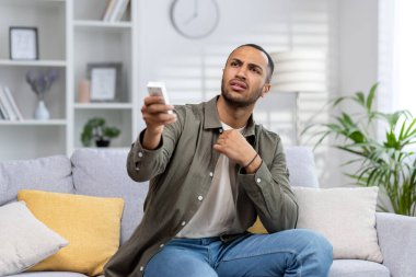 Müslüman genç adam evinde sıcaktan ve rahatsızlıktan muzdarip, elinde uzaktan kumandayla kanepede oturuyor ve klimayı açıyor..