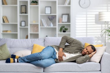 Genç Müslüman bir adam evinde kanepede yatıyor, hasta ve elleriyle karnını tutuyor, acı çekiyor, bulantı ve rahatsızlık hissediyor..