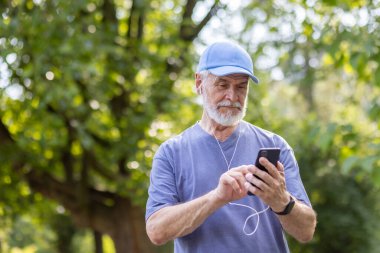 Yaşlı, kır saçlı, parkta spor yapan, kulaklık takan ve cep telefonu kullanan bir adam. Yakın plan fotoğraf..