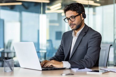 Genç bir Hintli işadamı ofiste dizüstü bilgisayarla çalışıyor ve kulaklık takıyor. Klavyeye konsantre yazma, sohbetler.