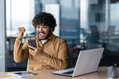 İş yerindeki İspanyol asıllı kazananın portresi, iş adamı online kazanç bildirimi aldı telefon uygulamasından, zaferi sevinçle kutlayan adam el ele tutuşarak, ofisin içinde dizüstü bilgisayarla çalışıyor..