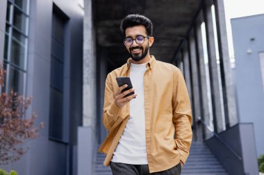 Genç, gülümseyen bir adam elinde bir telefonla, bir ofis binasının önünde yürüyor, mutlu bir şekilde akıllı telefondan bir uygulama kullanıyor, mesajları, tipleri okuyor ve internette geziniyor..