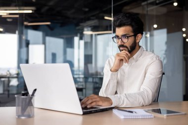 Genç Hintli bir adam ofiste dizüstü bilgisayarla çalışıyor, düşünceli ve ciddi bir şekilde ekrana bakıyor. Bilgi ve haber okuyor, bir e-posta aldı..
