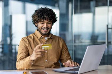 Genç Hintli bir adam ofiste dizüstü bilgisayarla çalışıyor ve elinde bir kredi kartı kullanıyor..