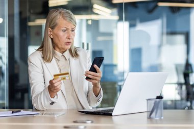 Confused senior businesswoman sitting at the desk in the office in a business suit, holding a credit card and looking worriedly at the phone screen. clipart