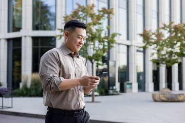 Asyalı genç ofis çalışanı iş yerinde telefonu kullanıyor, ofis binasının önünde duruyor, bir arama yapıyor, mesaj yazıyor..