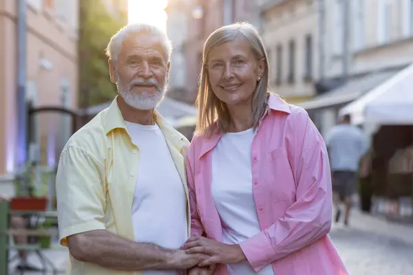 Portrait of an older couple, a man and a woman standing together on a city street, hugging and holding hands, smiling and posing for the camera.