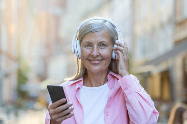 Portrait of an older beautiful gray-haired woman wearing headphones listening to music on the phone. Standing on a city street and smiling at the camera. Close-up photo.