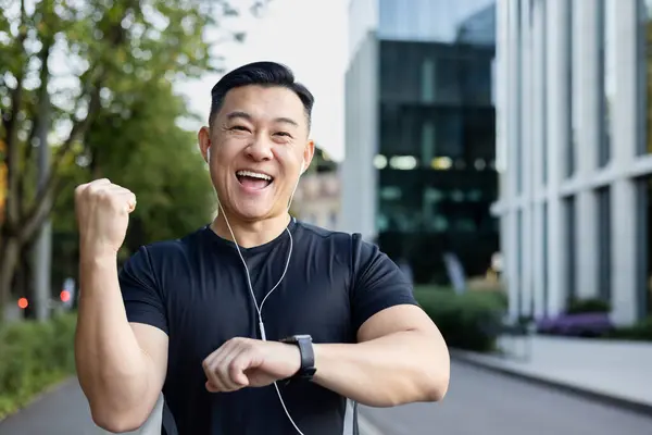 Portrait of a positive and happy young Asian male sportsman who is happy about the result of a run showing a smart watch, showing a success sign with his hand, smiling at the camera.