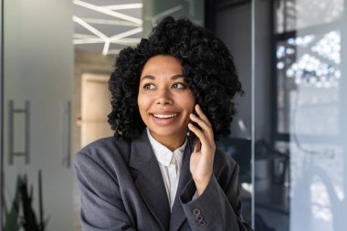 Ofiste takım elbiseli, gülümseyen ve telefonda konuşan güzel bir Afro-Amerikan kadının yakın plan portresi..