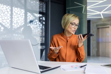 Kızgın kadın patron sesli mesaj kaydediyor, iş kadını ofis içinde bilgisayarla çalışmaktan rahatsız, akıllı telefondan online iletişim için başvuru yapıyor, mesaj bırakıyor. .