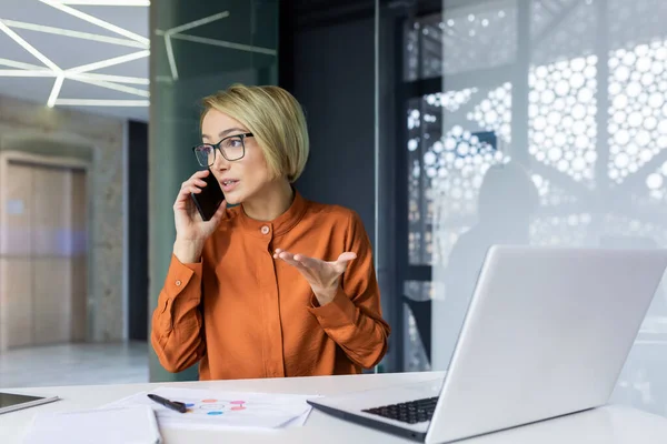 stock image Serious and angry female boss talking on the phone at workplace, business woman working with laptop.