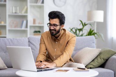 Genç Hintli erkek serbest bilgisayar kullanarak evde çalışıyor. Kanepeye güler yüzle oturur ve klavyeye yazar, karşılık verir, projeyi yürütür..