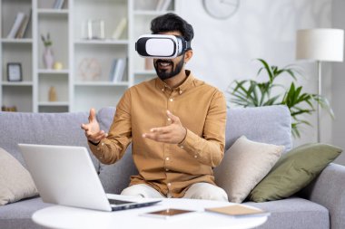 Hintli genç adam evinde kanepede dizüstü bilgisayarın önünde oturuyor, çalışıyor, çalışıyor ve sanal maskeyle konuşuyor..