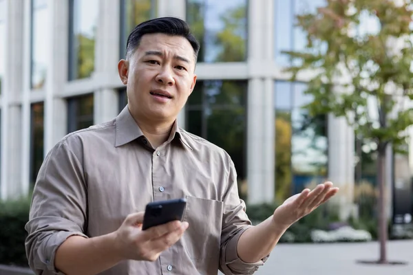 Close-up portrait of a sad young Asian man standing near an office building with a phone in his hands and looking worriedly at the camera, throwing up his hands in frustration.