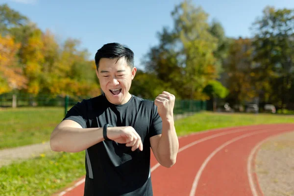 Close-up photo. Asian young male sportsman standing in the stadium and happy about the result at the finish line. He looks at the smart watch and makes a victory gesture with his hand.
