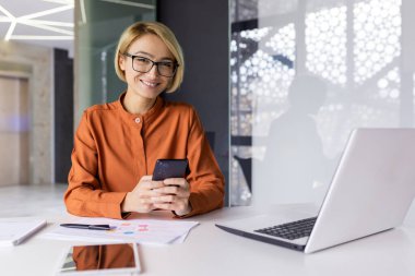 Ofisin içinde genç ve güzel bir kadının portresi, iş kadını gülümseyen ve kameraya bakan, telefondaki uygulamayı mutlu eden, masadaki dizüstü bilgisayarlı kadın..