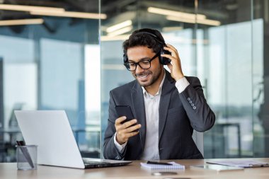 Genç, neşeli iş adamı ofiste dinleniyor, kulaklıklı adam müzik dinliyor, şarkı söylüyor ve dans ediyor, telefonda uygulama kullanıyor, elinde akıllı telefonuyla çalışıyor..