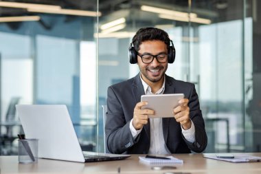 Ofis içinde kulaklıkları ve tabletle çalışan bir iş adamı çevrimiçi video izliyordu. Takım elbiseli bir adam iş yerinde takım elbisesiyle masasında mutlu bir şekilde oturuyordu..