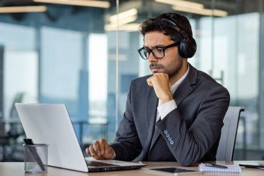 Ciddi düşünme ve ofis içinde dizüstü bilgisayarla çalışan, kulaklıklı, iş yerinde ses kitapları ve podcast dinleyen bir adam..