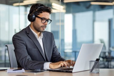 Ciddi düşünme ve ofis içinde dizüstü bilgisayarla çalışan, kulaklıklı, iş yerinde ses kitapları ve podcast dinleyen bir adam..