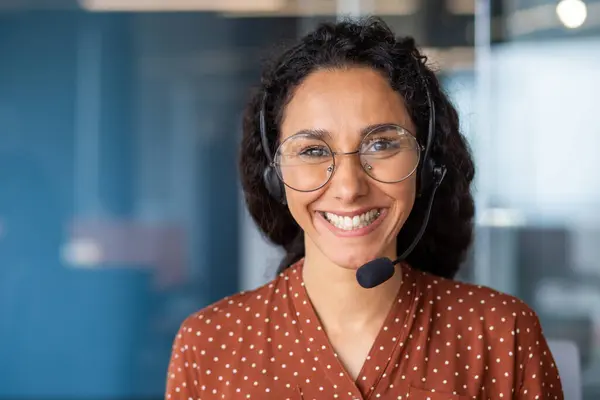 Close-up portrait of a woman with a headset, an online customer support worker smiling and looking at the camera, a business woman advising clients, working inside the office, using video call.