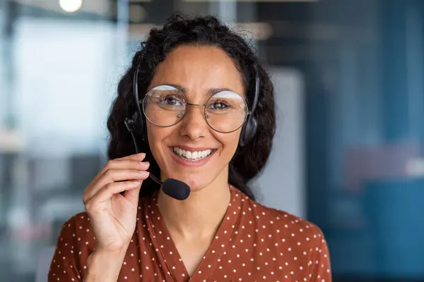 Close-up portrait of a woman with a headset, an online customer support worker smiling and looking at the camera, a business woman advising clients, working inside the office, using video call.