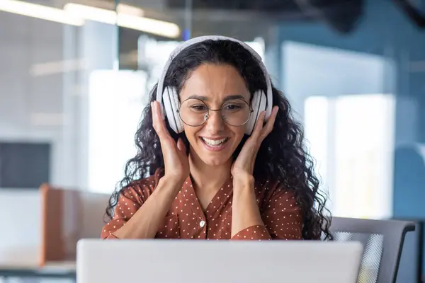 Young beautiful woman in headphones working with laptop inside office at workplace, joyful and satisfied business woman watching online webinar, educational video course, learning remotely.