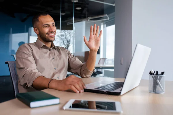 Happy successful and satisfied man talking on video call using laptop inside office, businessman waving hand smiling and greeting colleagues and partners, remote online meeting presentation.