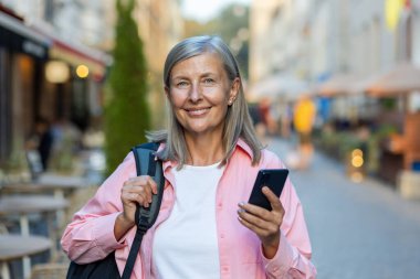 Caddenin ortasında sırt çantalı, cep telefonu kullanan, kameraya bakıp gülümseyen güzel bir kadının portresi..