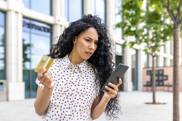 Upset and disappointed woman received money transfer error, Hispanic woman with bank credit card and phone cheated walking down street outside office building.